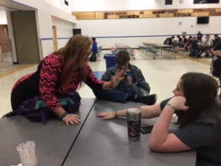 Three students at a table in a large room; one smacks the table.