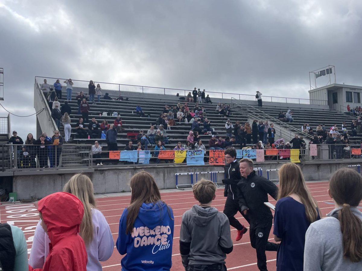 Two athletes sprint to the finish line at the end of their 4 x 100 meter relay, while middle school students work together to get their times.
